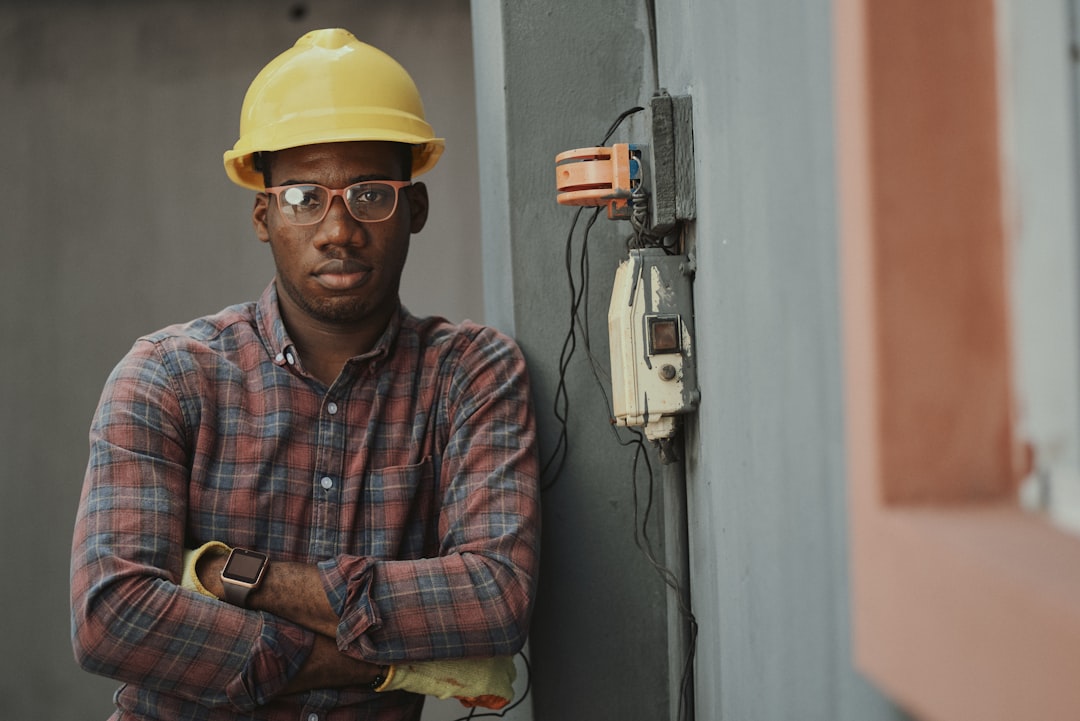 A person in a yellow hard hat and plaid shirt leaning against a wall